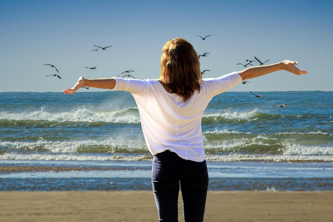 Woman on beach, arms out stretched out, birds in sky. EFT Tapping