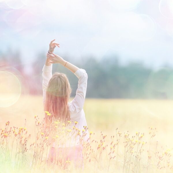 A young woman in a field in hazy sunshine, arms above head, depicting joyful feelings someone might feel after having hypnotherapy sessions